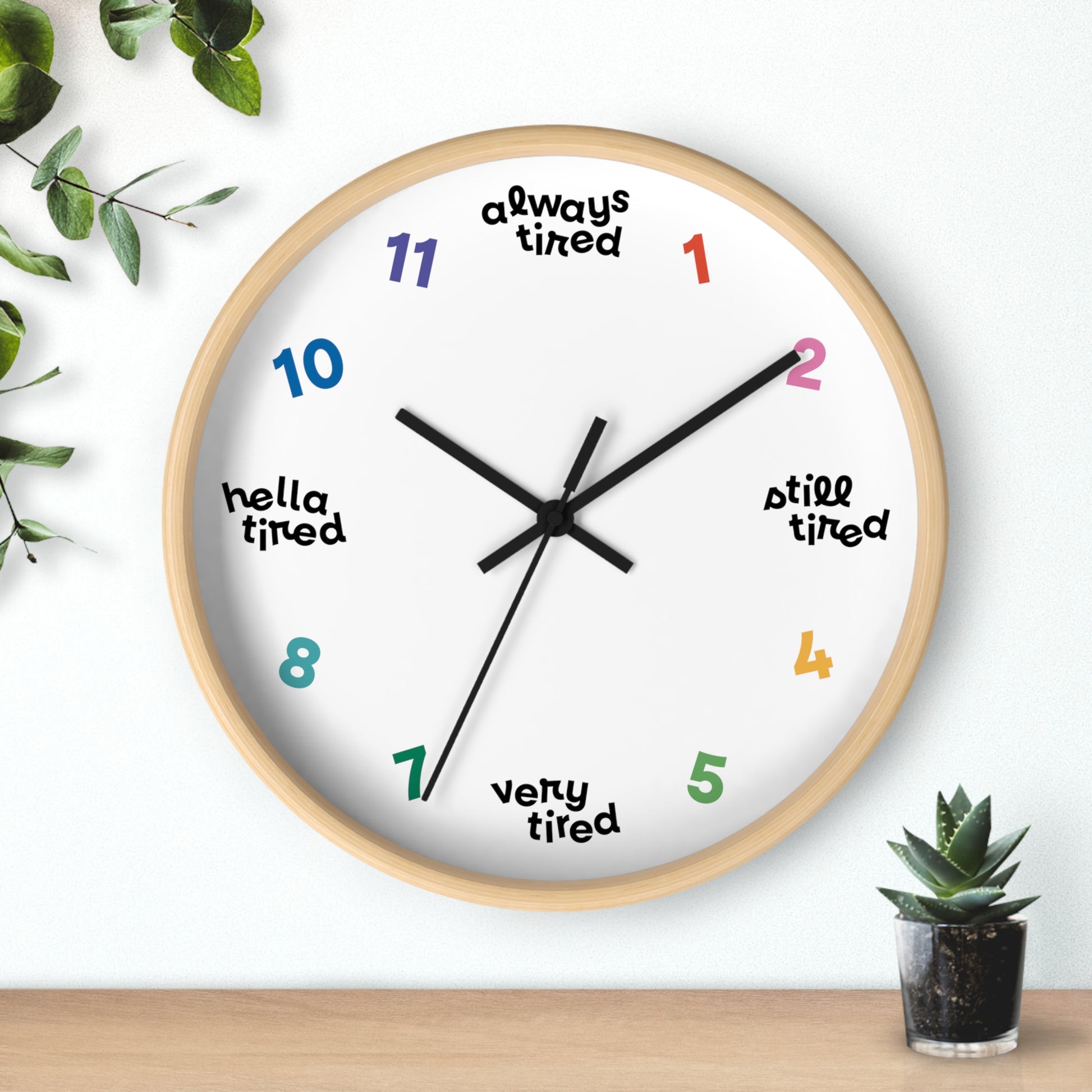 Hanging wall clock with a swedish minimal design. On 4 quadrants, there are variations of "tired" instead of the hour markers. The remaining numbers are cheerful bright colors. This clock has a wooden frame.