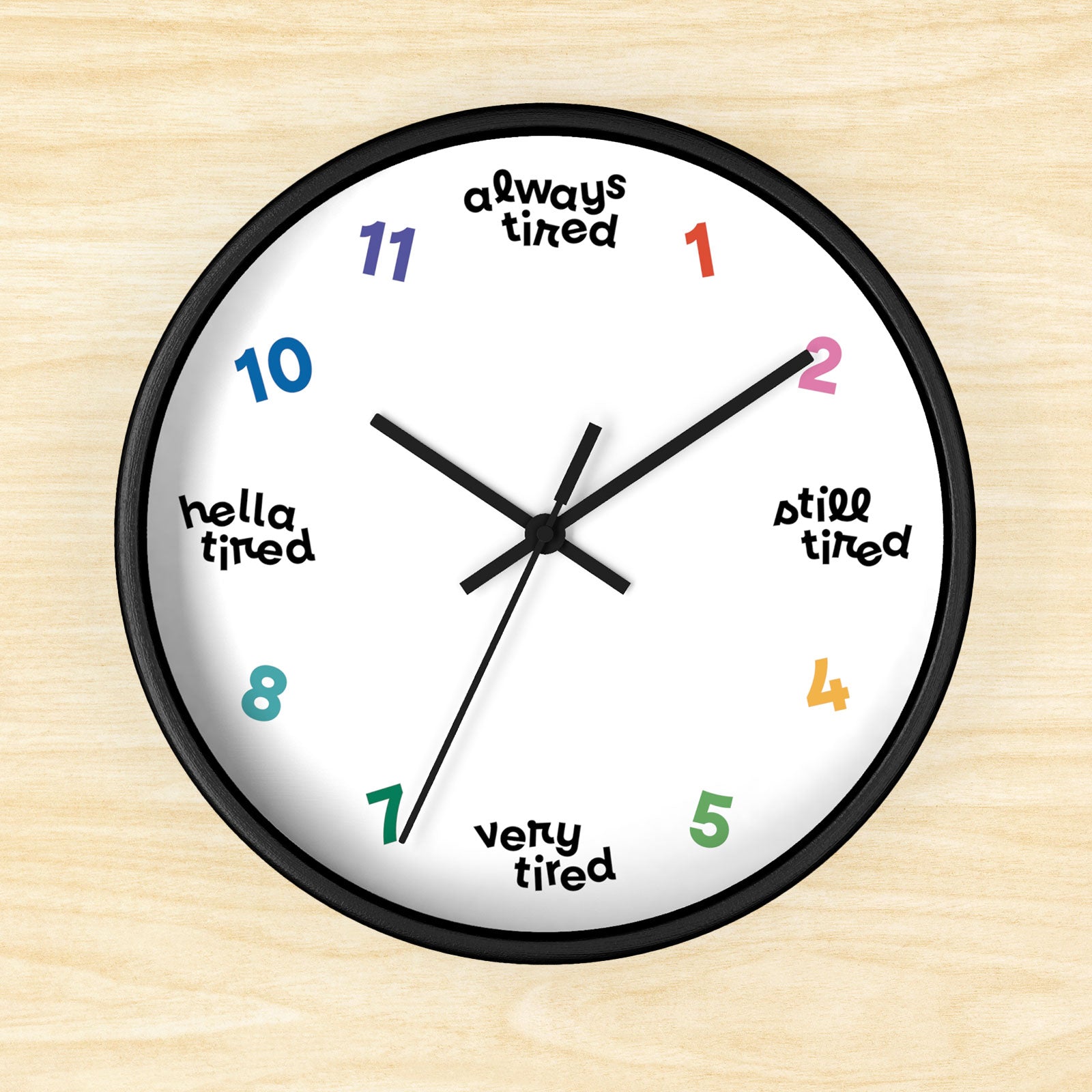 Hanging wall clock with a swedish minimal design. On 4 quadrants, there are variations of "tired" instead of the hour markers. The remaining numbers are cheerful bright colors. This clock has a black frame.